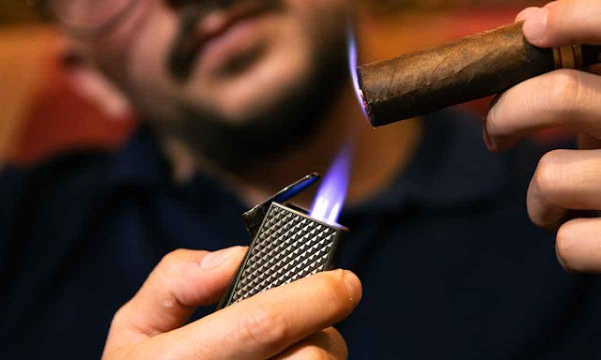 a bespectacled man lighting a cigar using a torch lighter